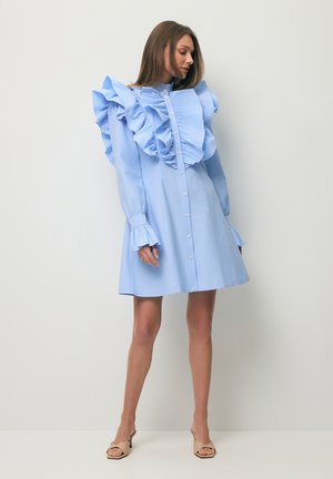 Vrouw draagt een lichtblauwe jurk met knopen, grote ruches op de schouders en borst, staat tegen een witte muur, beige open-toe hakken.