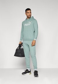 Tuta da uomo Puma in verde chiaro, composta da una felpa con cappuccio e logo e da pantaloni jogging abbinati. Include una borsa da viaggio nera e sneakers nere.