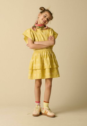 Jeune fille avec une coiffure en double chignon portant une robe jaune à motifs, une écharpe colorée, des chaussettes dépareillées et des baskets beige, souriante avec les bras croisés.