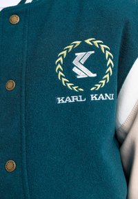 Turkusowa wełniana kurtka z beżowymi skórzanymi rękawami, z złotymi guzikami i haftowanym logo w laurowym wieńcu. Unikalny detal tekstury.