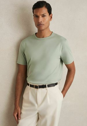 Tricou scurt de culoare verde mentă cu guler rotund, asociat cu pantaloni bej deschis și o curea neagră cu o cataramă aurie.