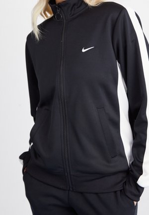 Person trägt eine schwarze Nike-Zip-Up-Jacke mit weißen Streifen an den Ärmeln und passende schwarze Hose, Hände in den Taschen.