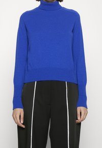 Kungsblå polo-tröja med ribbad textur och mudd, med en kortad design. Kombinerad med svarta byxor med vita sidodetaljer.