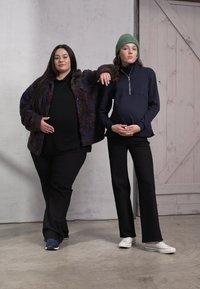 Duas pessoas grávidas estão lado a lado. Uma veste uma jaqueta felpuda colorida e calças pretas, enquanto a outra usa uma parte de cima escura com fecho e calças pretas.