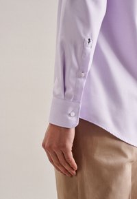 Camicia a maniche lunghe viola chiaro con colletto a punta e polsini abbottonati, caratterizzata da un dettaglio di ricamo floreale nero. Tessuto liscio.