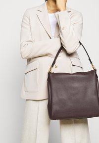 Person iført en beige blazer og hvide bukser, som holder en stor skindtaske i mørkebrown med tekstur og guldfarvet hardware på skulderen.