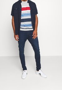 Avslappnad outfit med en marinblå kortärmad skjorta, grårandig t-shirt, mörka jeans och vita sneakers med svarta detaljer.