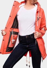 Orange vattentät jacka med ett vitt fleecefoder, dragkedjefickor, justerbar fåll och dragsko. Buren över en vit topp och mörka jeans.