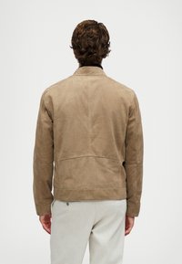 Άνδρας γυρισμένος προς τα πίσω, φορώντας ένα ανοιχτό καφέ σουέτ σακάκι και ανοιχτό γκρι παντελόνι, με φόντο έναν απλό λευκό τοίχο.