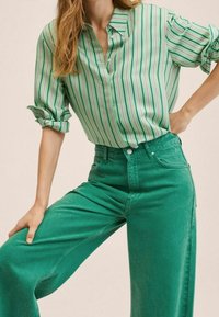 Blusa verde a righe con colletto, abbottonata a metà; abbinata a jeans verdi a vita alta e gamba larga con tasche frontali.