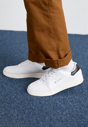 Hvide sneakers med en glat syntetisk overdel, grå hælaccent og lys gummisål. Båret med brune bukser med opsmøg og hvide sokker.