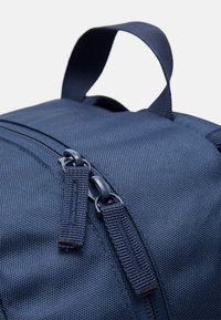 Sac à dos bleu marine avec un tissu texturé, des tirettes de fermeture à glissière doubles et une poignée de transport courte. Gros plan sur la zone de la fermeture et le matériau.