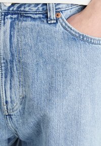 Jean en denim bleu clair avec une surface texturée, fermeture par bouton supérieur et une poche latérale. Présente des coutures visibles et une coupe classique.