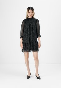 Mulher jovem em pé, vestindo um vestido mini de manga longa preto transparente com sutis riscas de zebra e sapatos slingback pretos de bico fino.