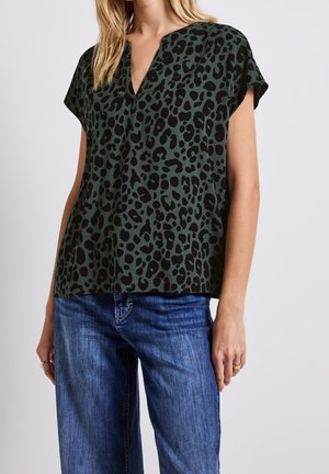 Vrouw draagt een donkergroene top met korte mouwen, zwarte luipaardvlekken en blauwe spijkerbroek, voor een effen lichte achtergrond.