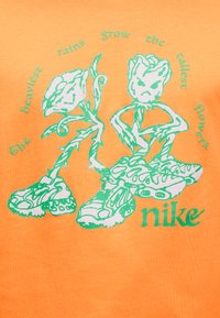 Πορτοκαλί μπλουζάκι με πράσινο γραφικό σχέδιο δύο στυλιζαρισμένων λουλουδιών με πρόσωπα, με τη λέξη "Nike" στο κάτω μέρος.