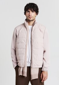 Helles rosa Daunenjacke mit Stehkragen, gestepptem Design und gerippten Strickakzenten am Saum. Getragen über einem weißen Hemd.