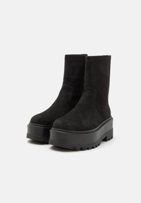 Par de botines de ante negro con suelas gruesas y plataforma, y suelas con tacos, sobre un fondo claro y sencillo.