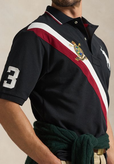 Mann iført svart pikéskjorte med hvite og røde diagonale striper, brodert emblemat, nummer 3 på ermet, og grønn genser knyttet rundt livet.