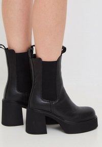 Botines de cuero negro hasta el tobillo con paneles elásticos en los laterales. Tacón robusto y punta redondeada, con una textura suave y un diseño minimalista.