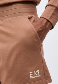 Το χέρι ενός ατόμου στην τσέπη καφέ σορτς Emporio Armani EA7 με μαζεμένη μέση και ραμμένο λογότυπο κοντά στο στρίφωμα.