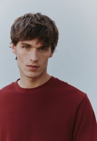 Rød struktureret sweatshirt med rund hals. Stoffet er ribbet, tætsiddende ved halsen, med en glat drapering. Modellen har uordentlig mørkt hår og en neutral baggrund.