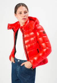 Vrouw in een glanzende rode pufferjas met capuchon, witte shirt en blauwe jeans, die iets naar de zijkant kijkt met haar handen in haar zakken.