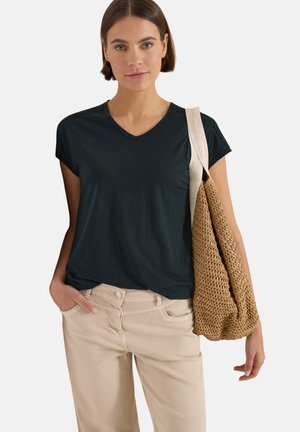 Femme portant un t-shirt noir à col en V et un pantalon beige, portant un sac cabas tressé marron avec une large bandoulière beige sur l’épaule.