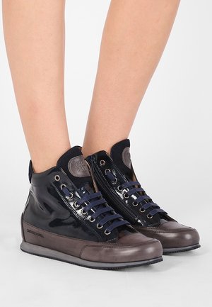 Nogi w czarno-brązowych wysokich trampkach z granatowymi sznurówkami i bocznymi zamkami błyskawicznymi na szarej powierzchni z białym tłem.