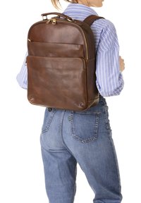 Sac à dos en cuir marron au design structuré, comportant deux poches avant et des zips dorés. Texture lisse, accents minimalistes.