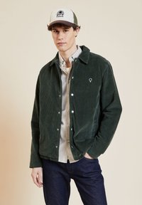 Veste en velours côtelé verte avec fermeture à boutons-pression, ornée d'un petit logo sur la poitrine, associée à une chemise grise à boutons et un jean foncé.