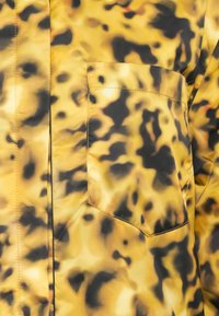 Tessuto giallo e nero a motivo con una texture lucida; presenta una tasca sul petto. Il design include macchie e vortici astratti.