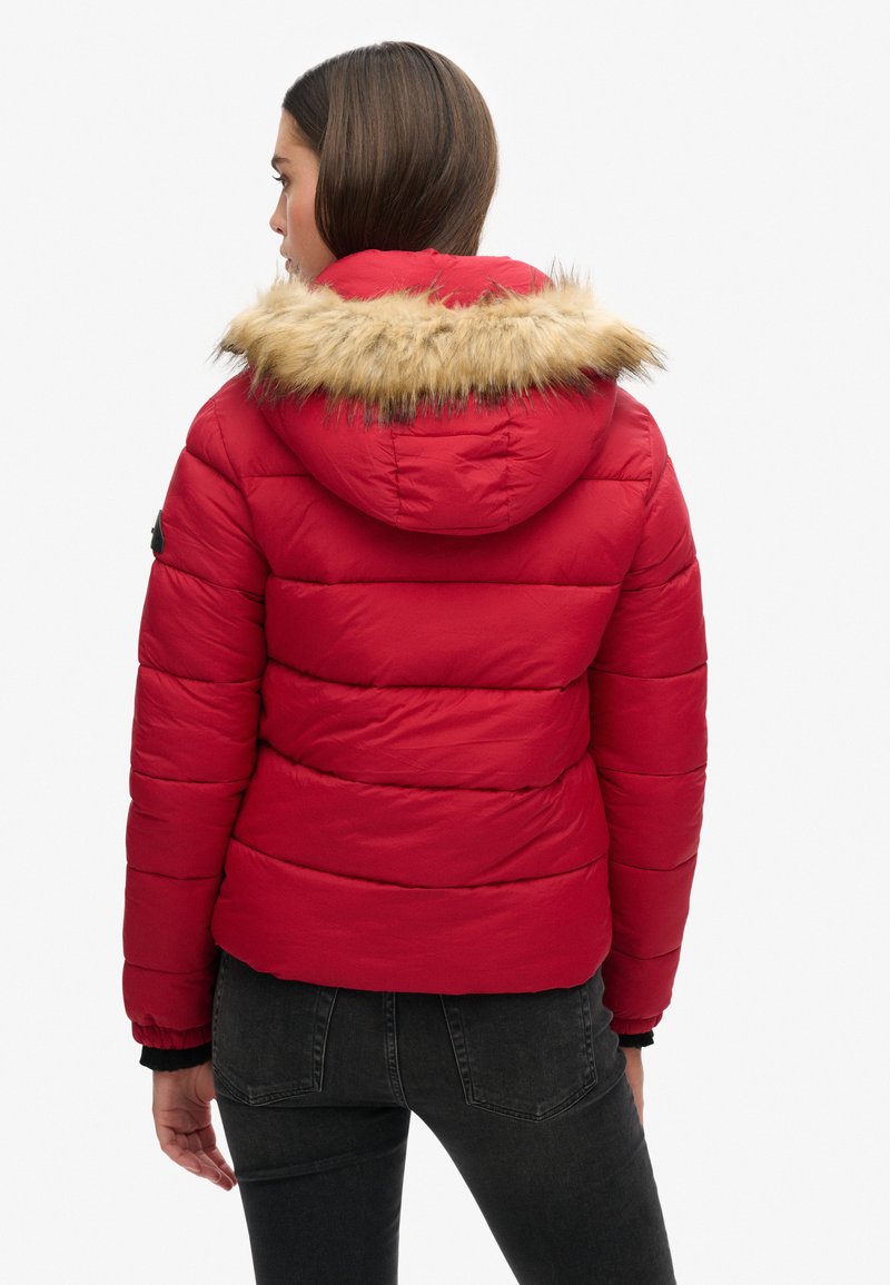 Giacca a vento rossa con cappuccio foderato in pelliccia sintetica, design trapuntato, polsini elasticizzati e vita aderente, realizzata in materiale leggero.