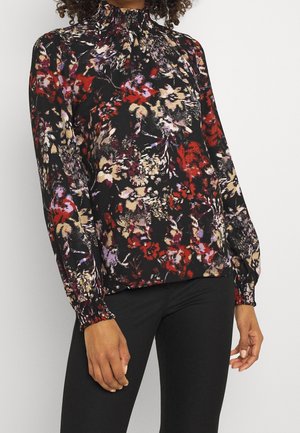 Femme portant un chemisier fleuri à manches longues avec un col haut froncé et un pantalon noir, debout devant un fond uni.