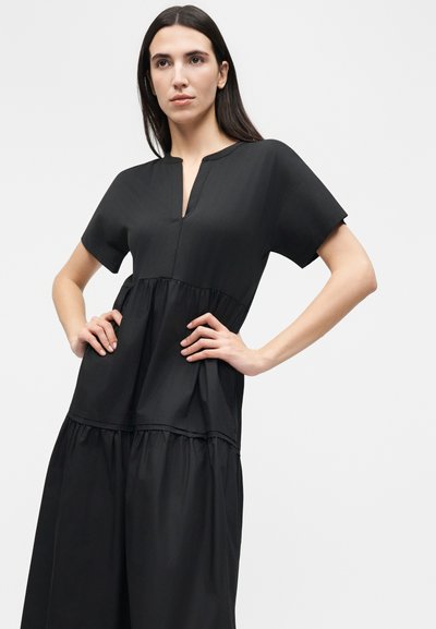 Γυναίκα με μακριά σκούρα μαλλιά που φοράει μαύρο φόρεμα με κοντά μανίκια και V-λαιμόκοψη, στέκεται με τα χέρια στη μέση μπροστά σε ένα απλό φωτεινό φόντο.