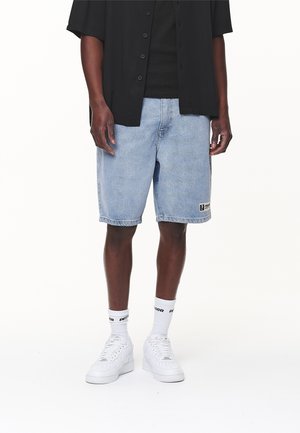 Jasnoniebieskie szorty dżinsowe o luźnym kroju, do kolan, z naszywką logo w kontrastowym kolorze. Noszone z białymi trampkami i skarpetkami załóżonymi do kostki.