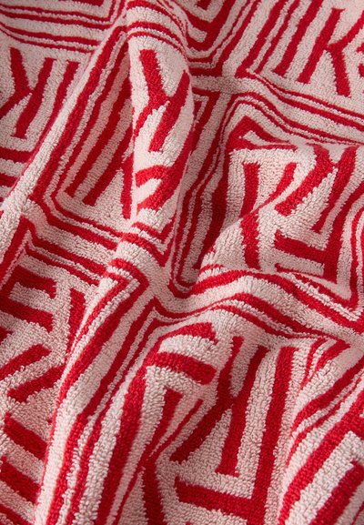 Κοντινή εικόνα υφασμάτινης επιφάνειας με ανάγλυφα κόκκινα γεωμετρικά γράμματα σε ανοιχτό ροζ φόντο, που δείχνει πτυχές και μαλακούς βρόγχους.