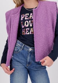 Purple, textured, sleeveless vest with a rounded collar over a dark shirt with "PEACE LOVE HEART" in colorful letters. Worn with blue jeans.