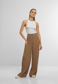 Lichtbruine, geplooide wijde broek gecombineerd met een strak wit tanktop. Model staat in sneakers tegen een neutrale achtergrond.