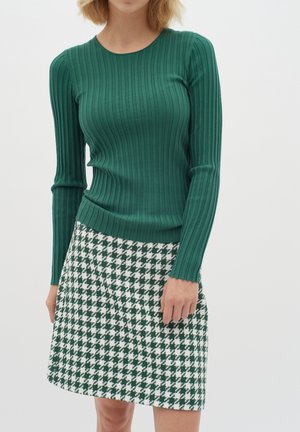 Žalias juostuotas ilgų rankovių megztinis derinamas su žaliu ir baltu houndstooth rašalo trumpu sijonu. Abu drabužiai pasižymi prigludusia forma.