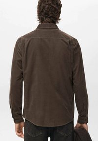 Camisa de pana marrón de manga larga, con puños abotonados y un cuello clásico. Presenta un dobladillo curvado y una textura suave.