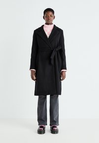 Black wool coat with a belt, lapel collar, and long sleeves. Paired with a pink striped sweater and grey jeans, and glossy platform shoes.
