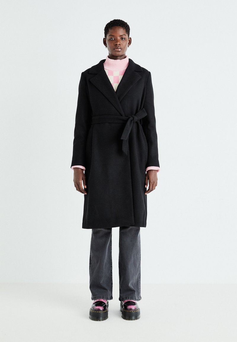 Black wool coat with a belt, lapel collar, and long sleeves. Paired with a pink striped sweater and grey jeans, and glossy platform shoes.