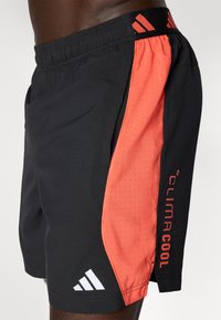 Gros plan sur une personne portant un short de sport noir avec un panneau latéral rouge comportant de petites perforations et le logo "Climacool".