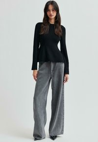 En ung kvinna står klädd i en tajt svart tröja och vida, urtvättade grå jeans mot en ljus neutral bakgrund.