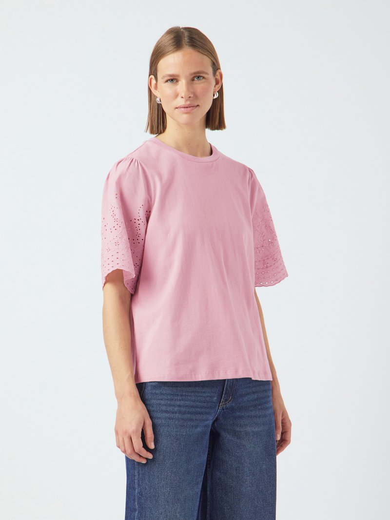 T-shirt en coton rose à manches courtes et volumineuses avec broderie en œillet. Associé à un jean large bleu, sur fond uni.