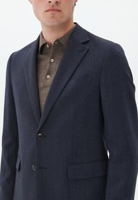 Marineblauer, maßgeschneiderter Blazer aus strukturiertem Stoff, mit einem Ein-Knopf-Verschluss und einer linken Brusttasche, kombiniert mit einem braunen Hemd mit Kragen.