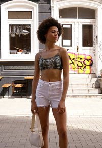 Top cropped com padrão de pele de cobra, com alças finas, e calções de ganga brancos. Modelo segura uma pequena bolsa de creme, em pé numa rua pavimentada.