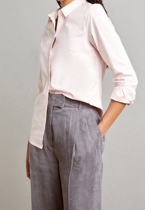 Kvinde iført lyserød skjorte med knapper og op-rullede ærmer samt højtaljede grå plisserede bukser, med hånden i lommen, foran ensfarvet baggrund.