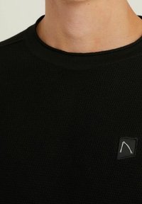 Czarna teksturowana bluza z okrągłym dekoltem i małą naszywką z logo na piersi, podkreślająca prosty i funkcjonalny design.